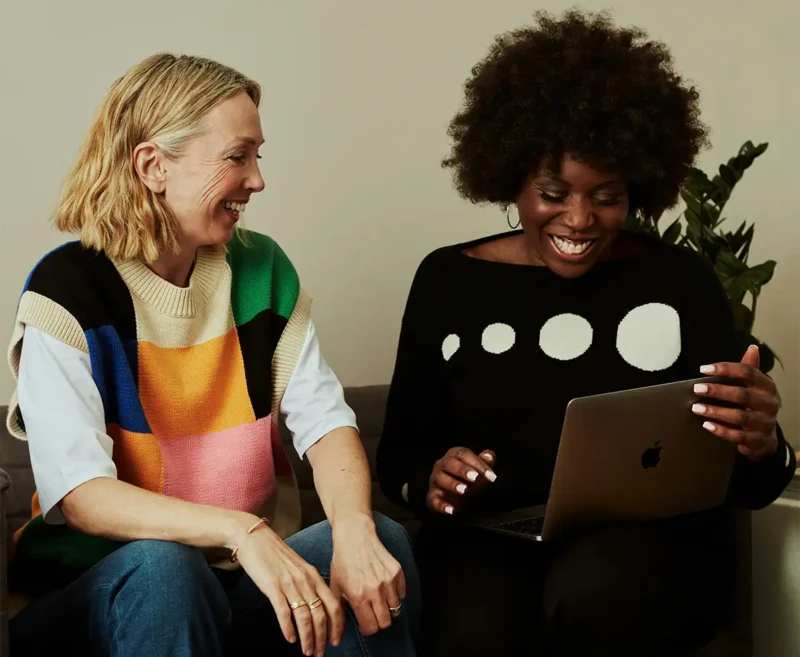 two women sitting on a couch smiling and one is-looking at a laptop